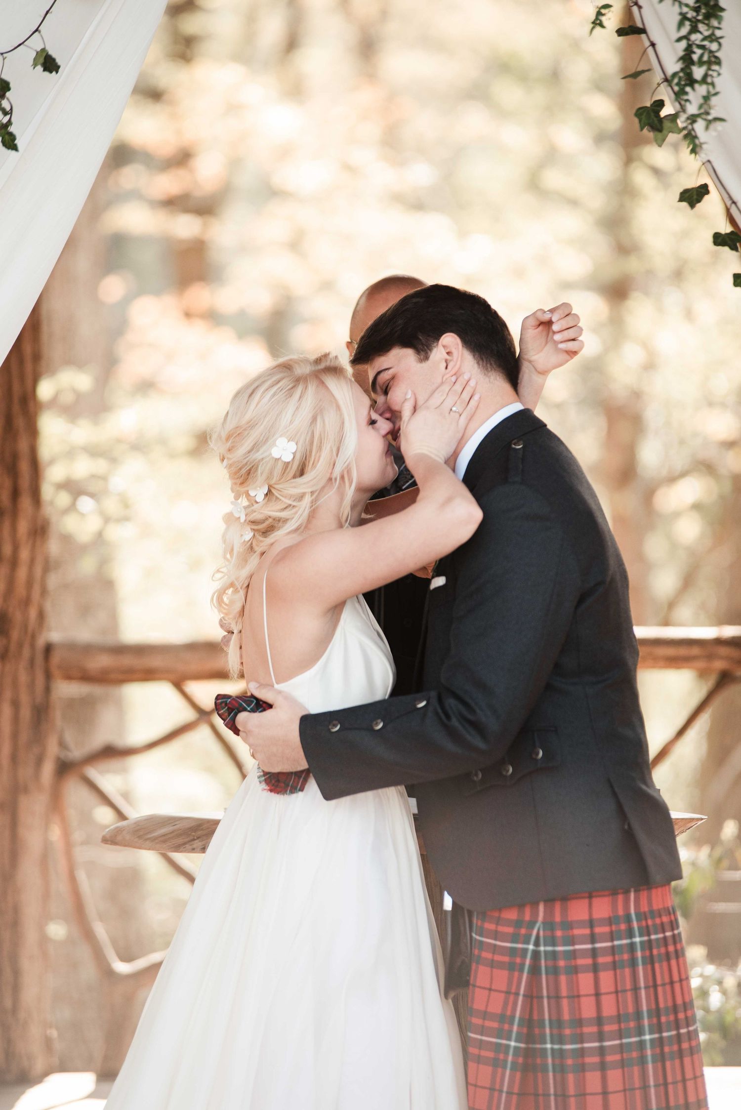 Bride and groom share first kiss at outdoor woodland wedding ceremony, groom wearing traditional Scottish tartan kilt.