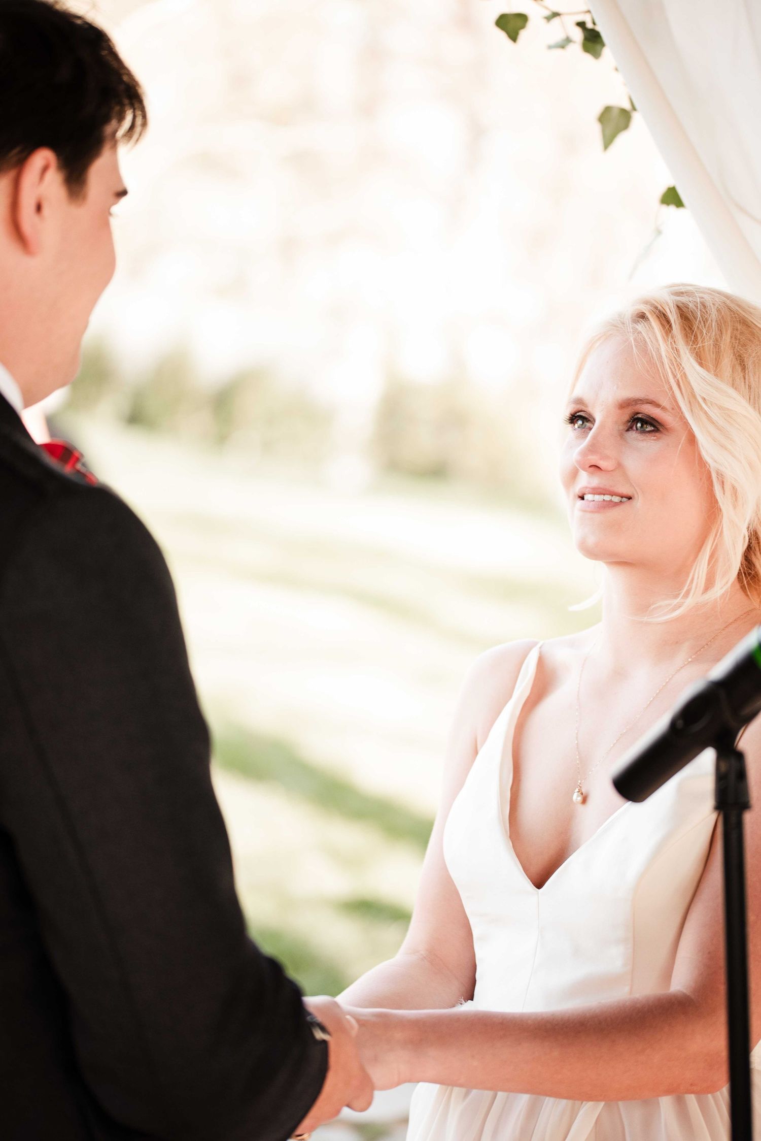 Bride smiling at groom during outdoor wedding ceremony, holding hands under a white draped arch.