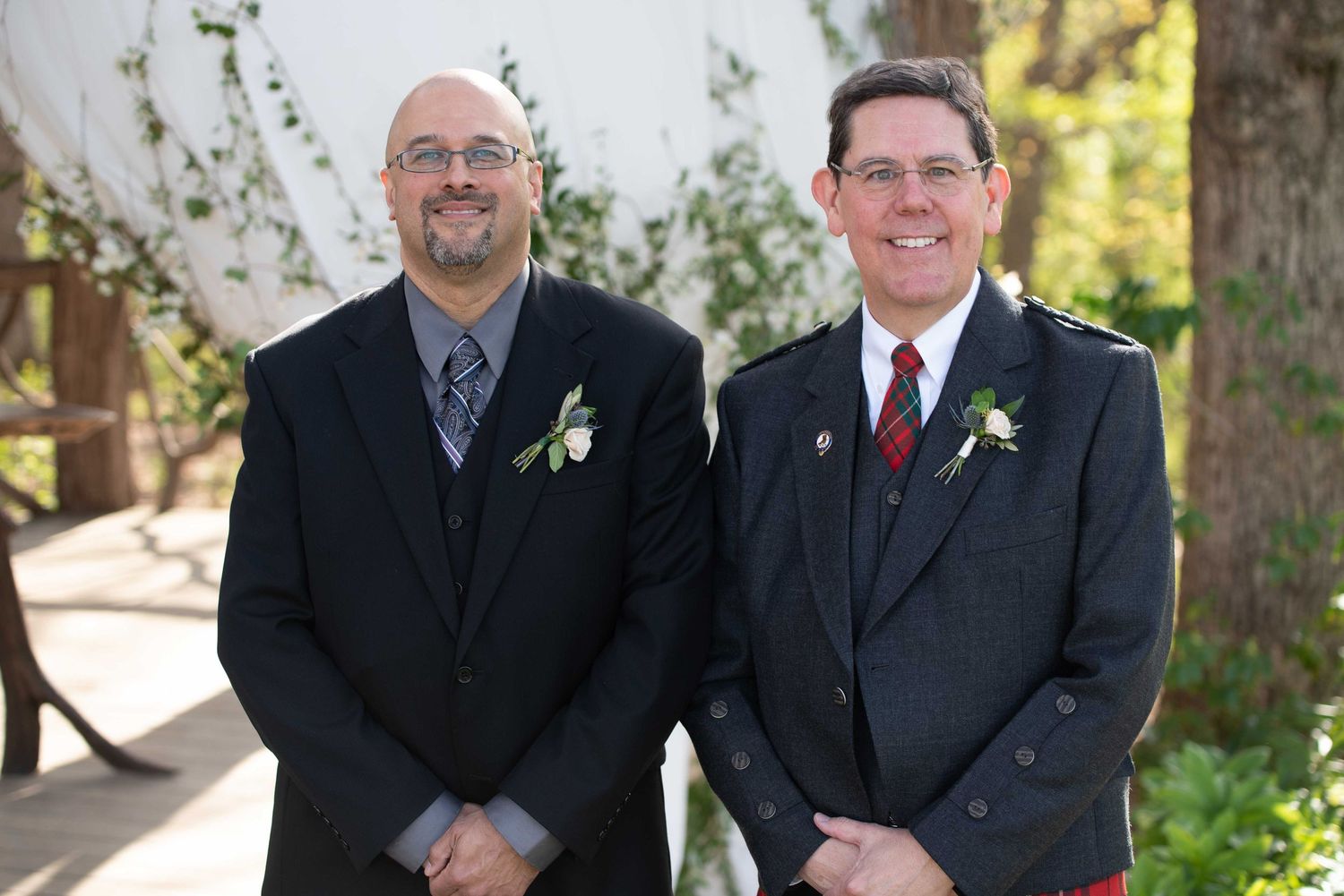 Two men in formal attire with boutonnieres pose outdoors at a wedding, one in a suit and one in a tartan kilt jacket.