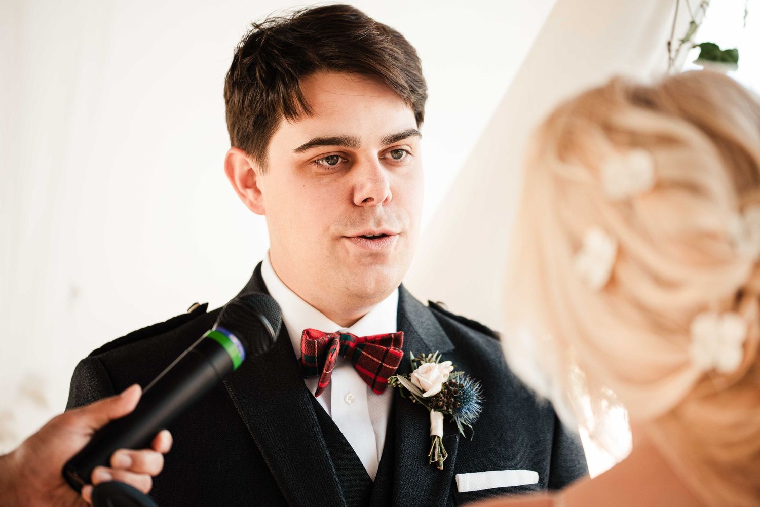 Groom in black suit with red bow tie and boutonniere reciting wedding vows to blonde bride.