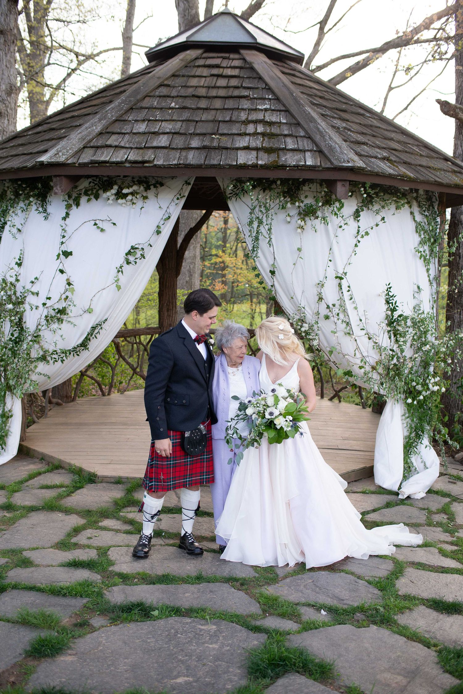 Bride and groom in Scottish kilt stand with elderly guest under decorated gazebo with white draping and greenery.