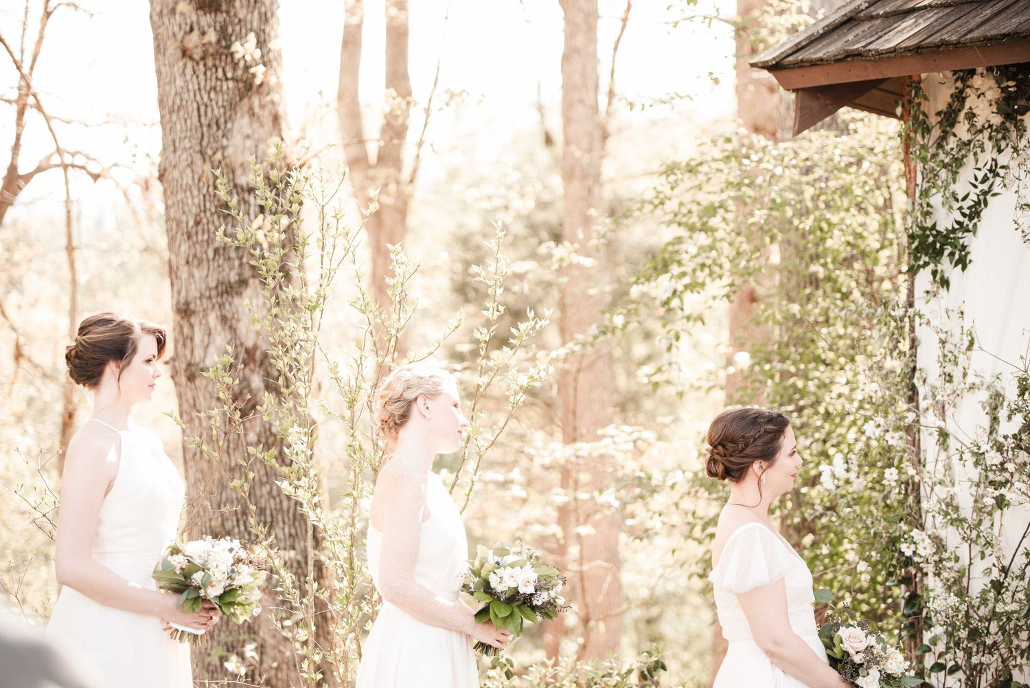 Three brides in white gowns holding bouquets stand among blooming trees at an outdoor woodland wedding ceremony.