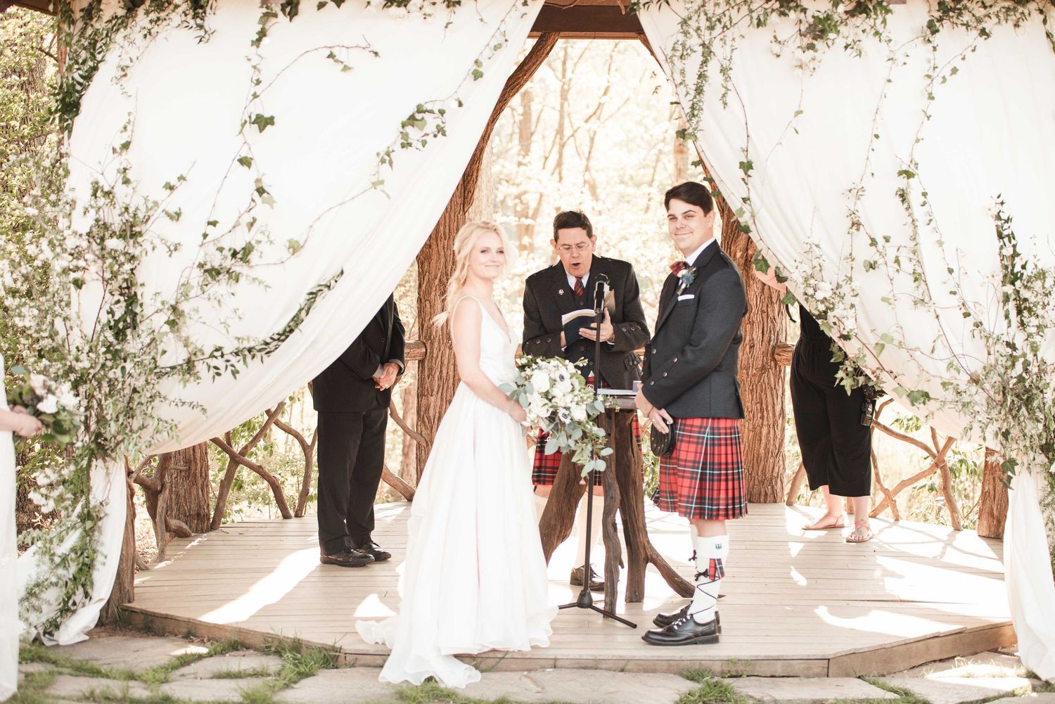 Bride and groom stand at floral altar during outdoor wedding ceremony, groom wearing traditional Scottish kilt.