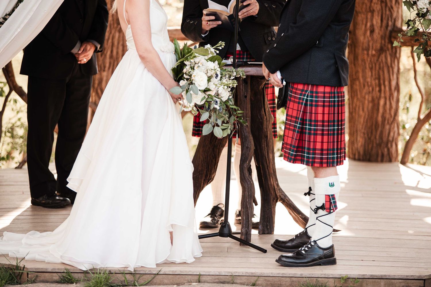 Wedding ceremony with bride in white gown holding bouquet and groom in red tartan kilt standing at rustic altar.
