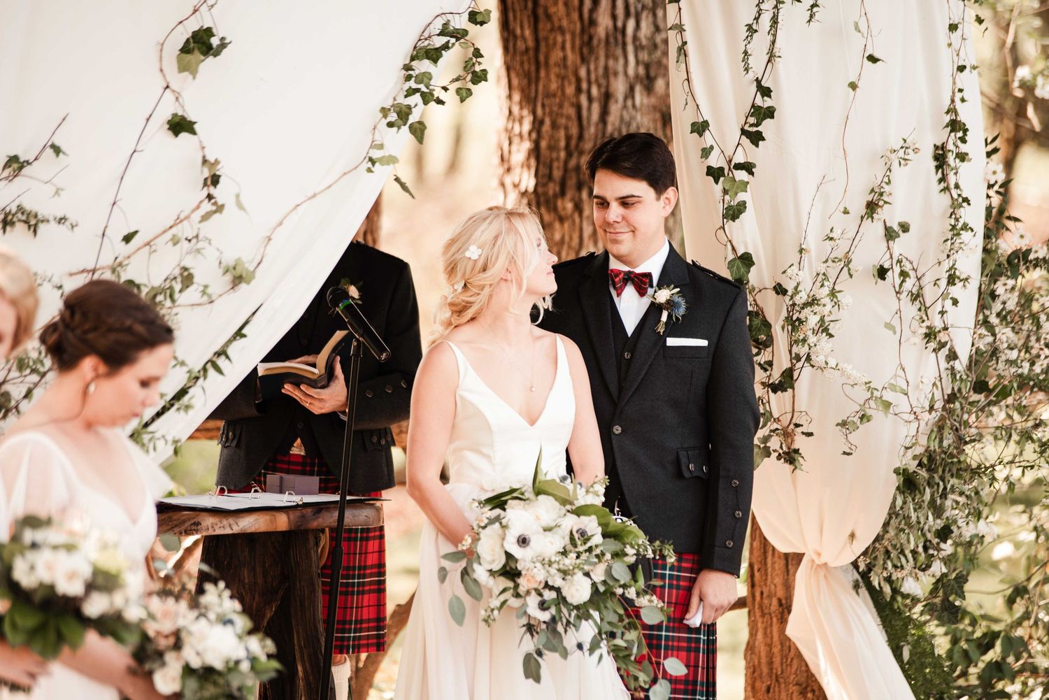 Bride and groom stand at floral wedding altar, groom in kilt and bow tie, bride holding white bouquet.