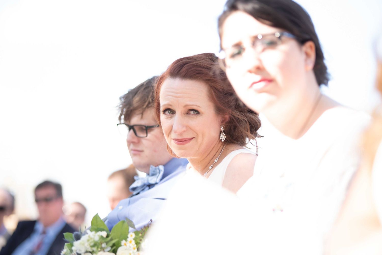 Emotional bride holding white bouquet with guests seated behind her at an outdoor wedding ceremony.
