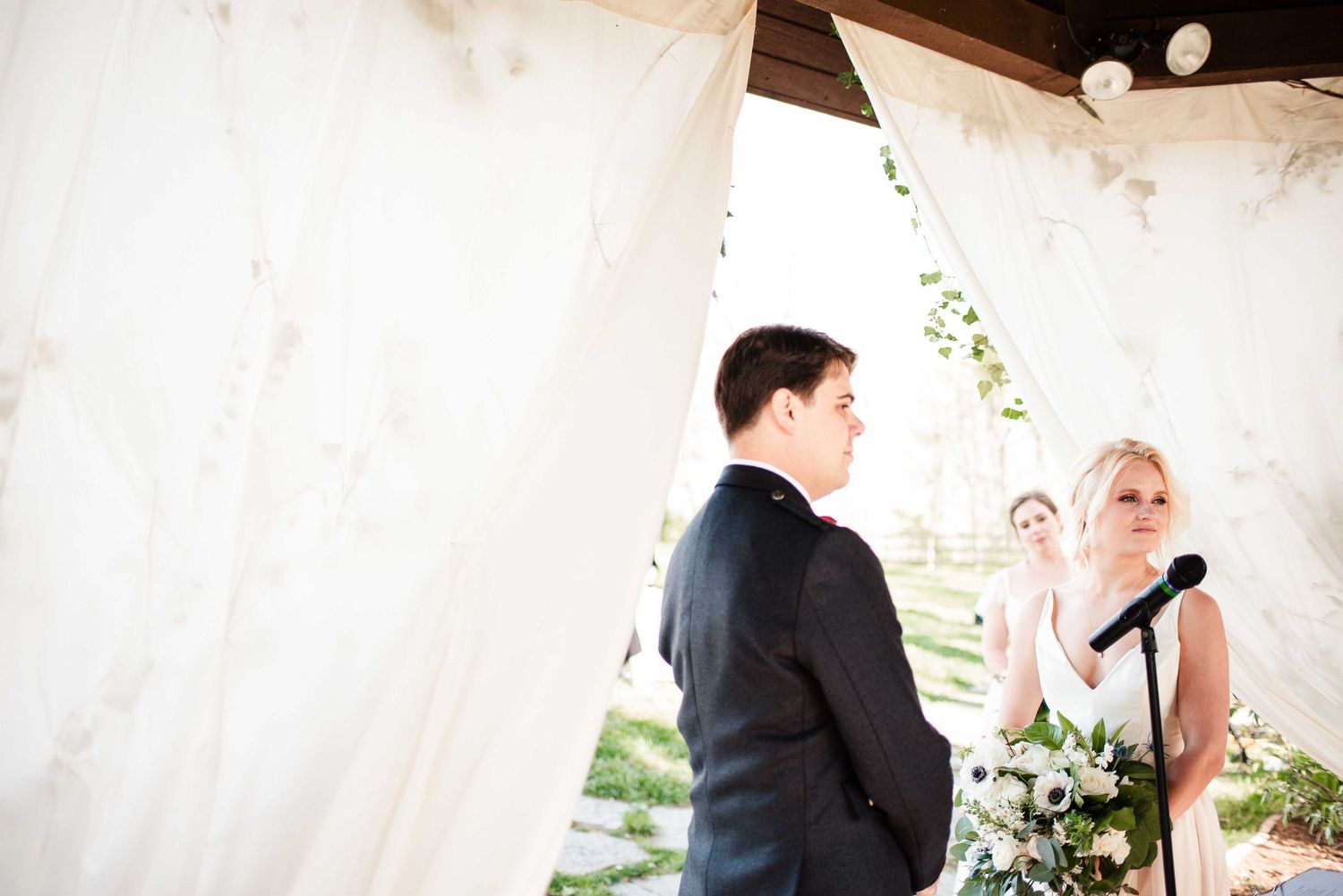 Bride and groom exchange vows under a white draped wedding arch during an outdoor ceremony.