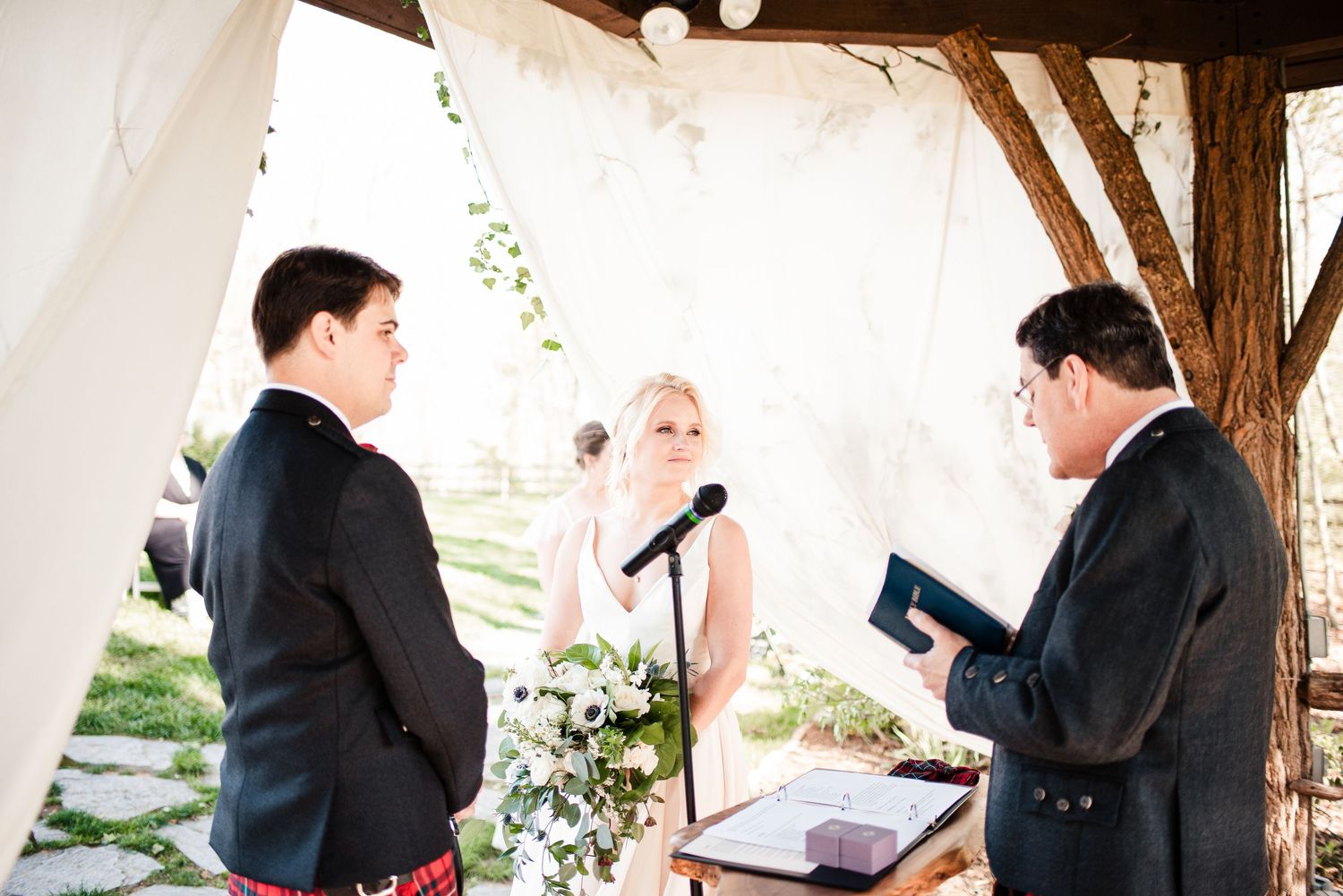 Bride and groom exchange vows at outdoor wedding ceremony with officiant holding book under rustic wooden pergola.