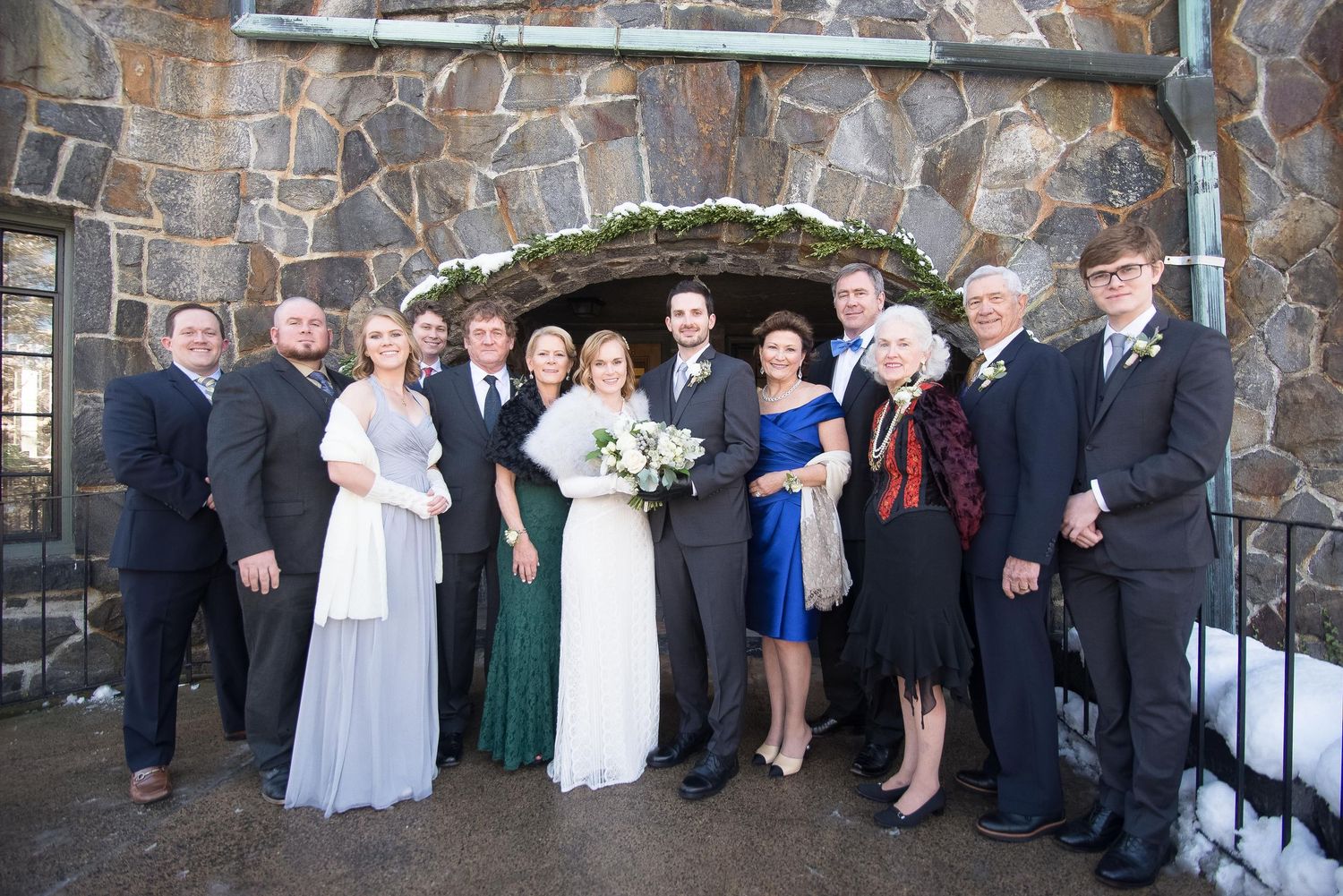 Wedding party poses in formal attire before a stone arch backdrop outdoors in winter.