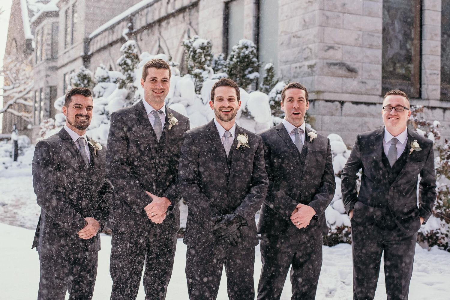 Five groomsmen in dark suits pose together outdoors in falling snow during a winter wedding celebration.