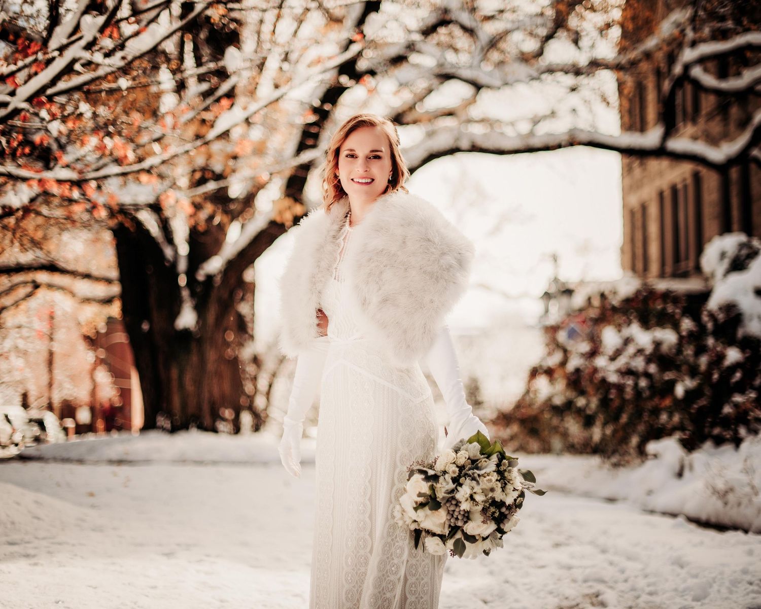 Smiling bride in white gown and fur stole holding bouquet in snowy winter outdoor setting.
