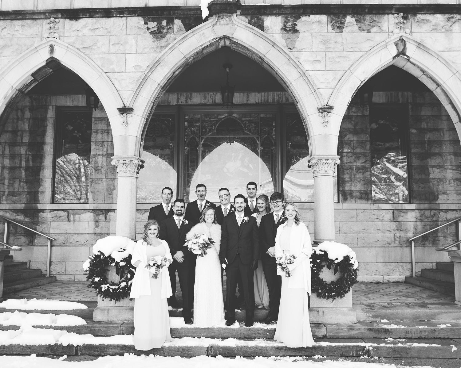 Black and white winter wedding party posed on snowy church steps with Gothic arched stone entrance and holiday wreaths.