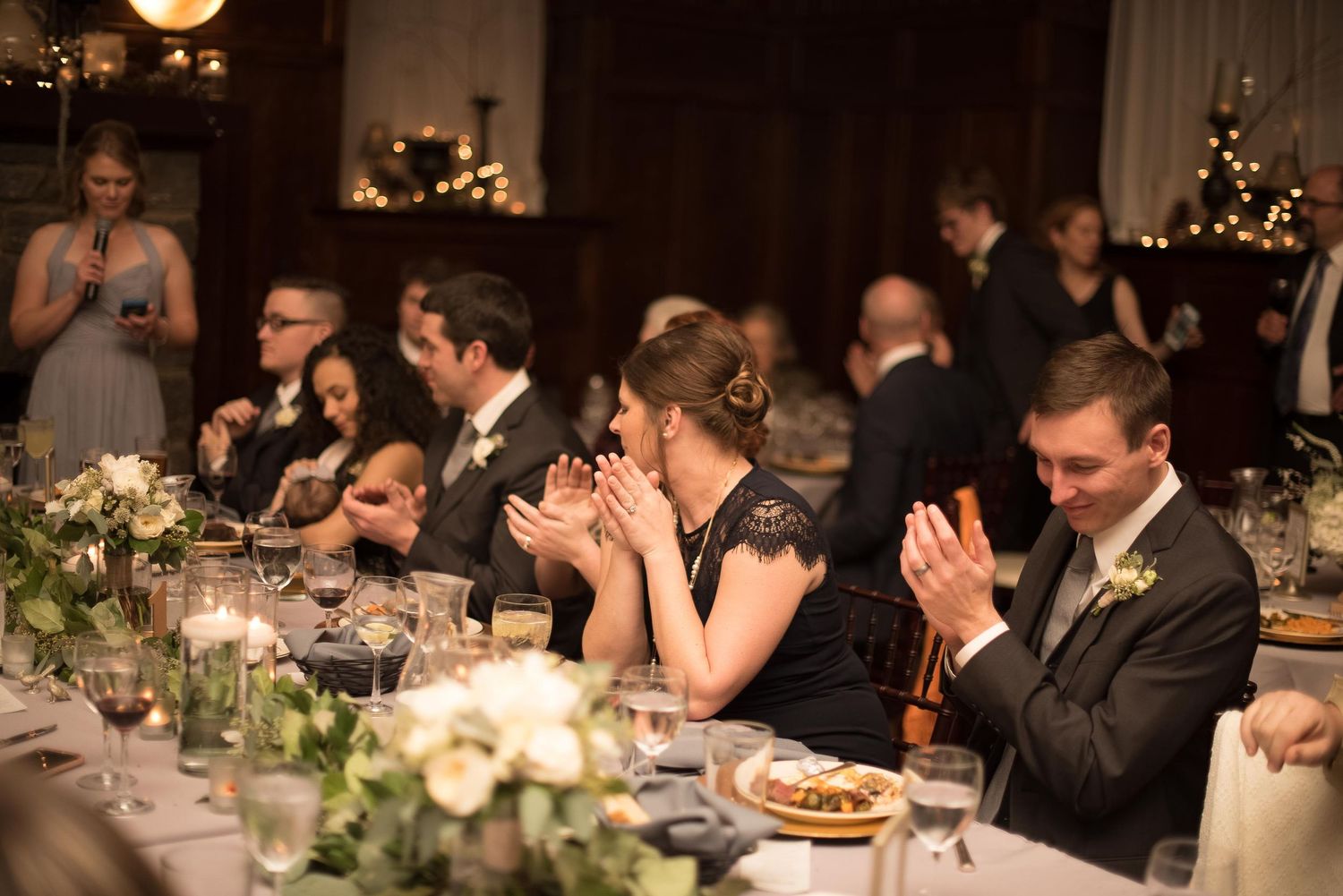Guests applaud and kiss at an elegant wedding reception dinner table with candles and white floral centerpieces.