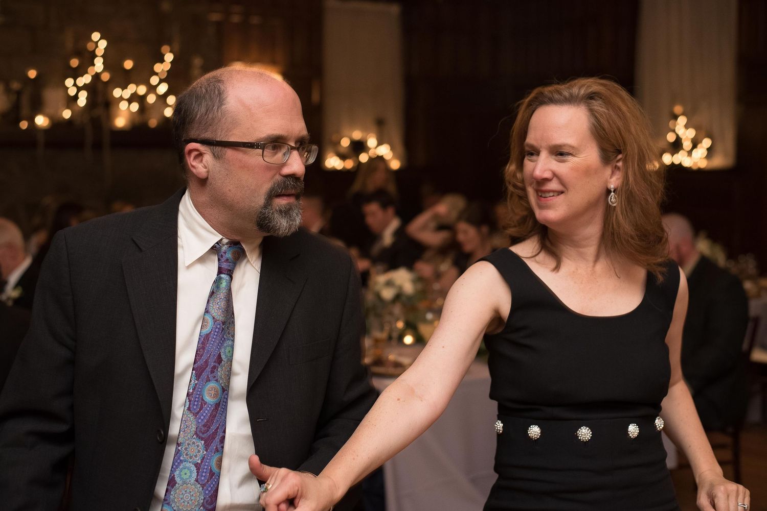 Couple holding hands and smiling at elegant indoor event, man in suit and woman in black dress.