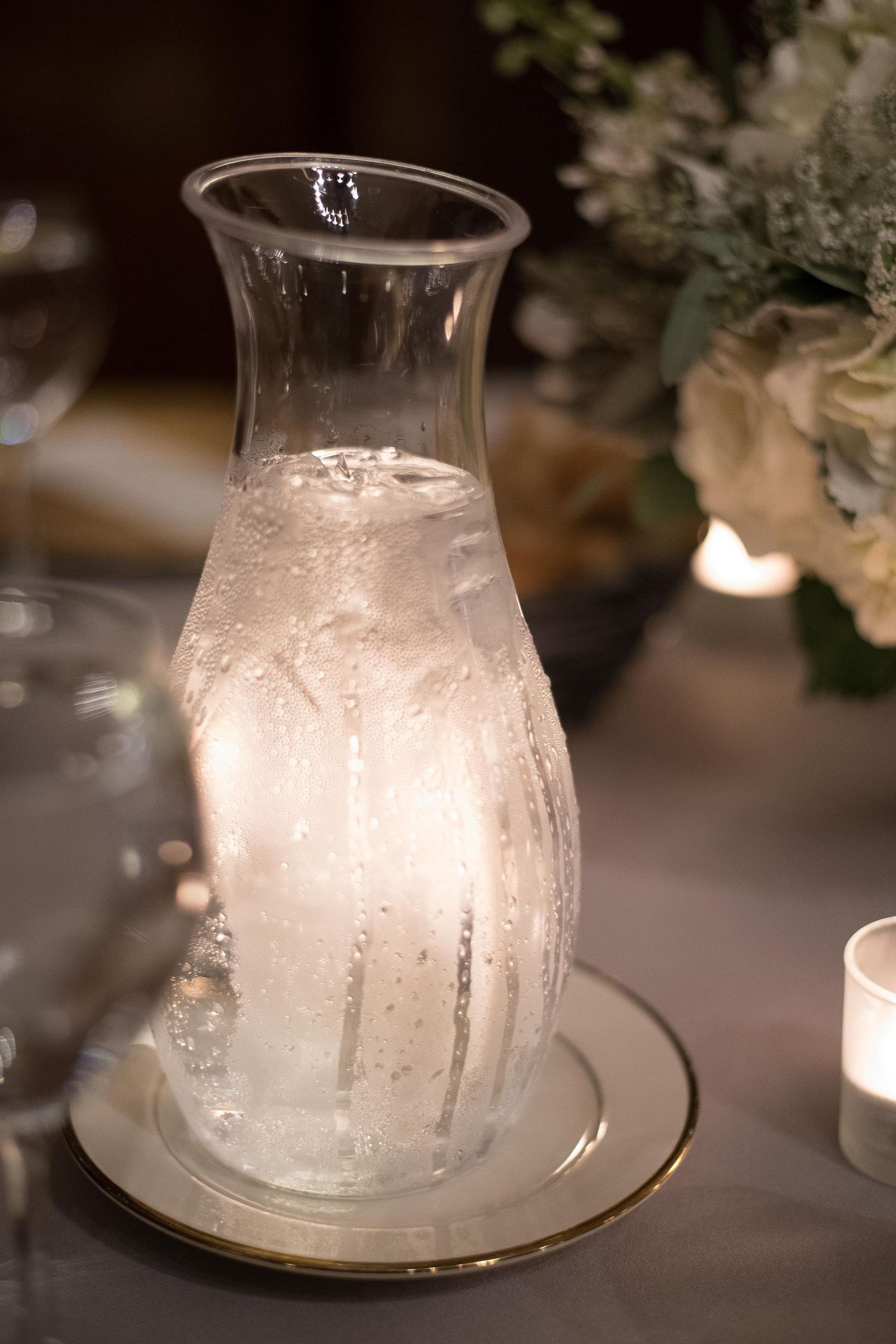Glass carafe filled with ice water on a gold-rimmed plate, glowing softly at an elegant candlelit dinner table.