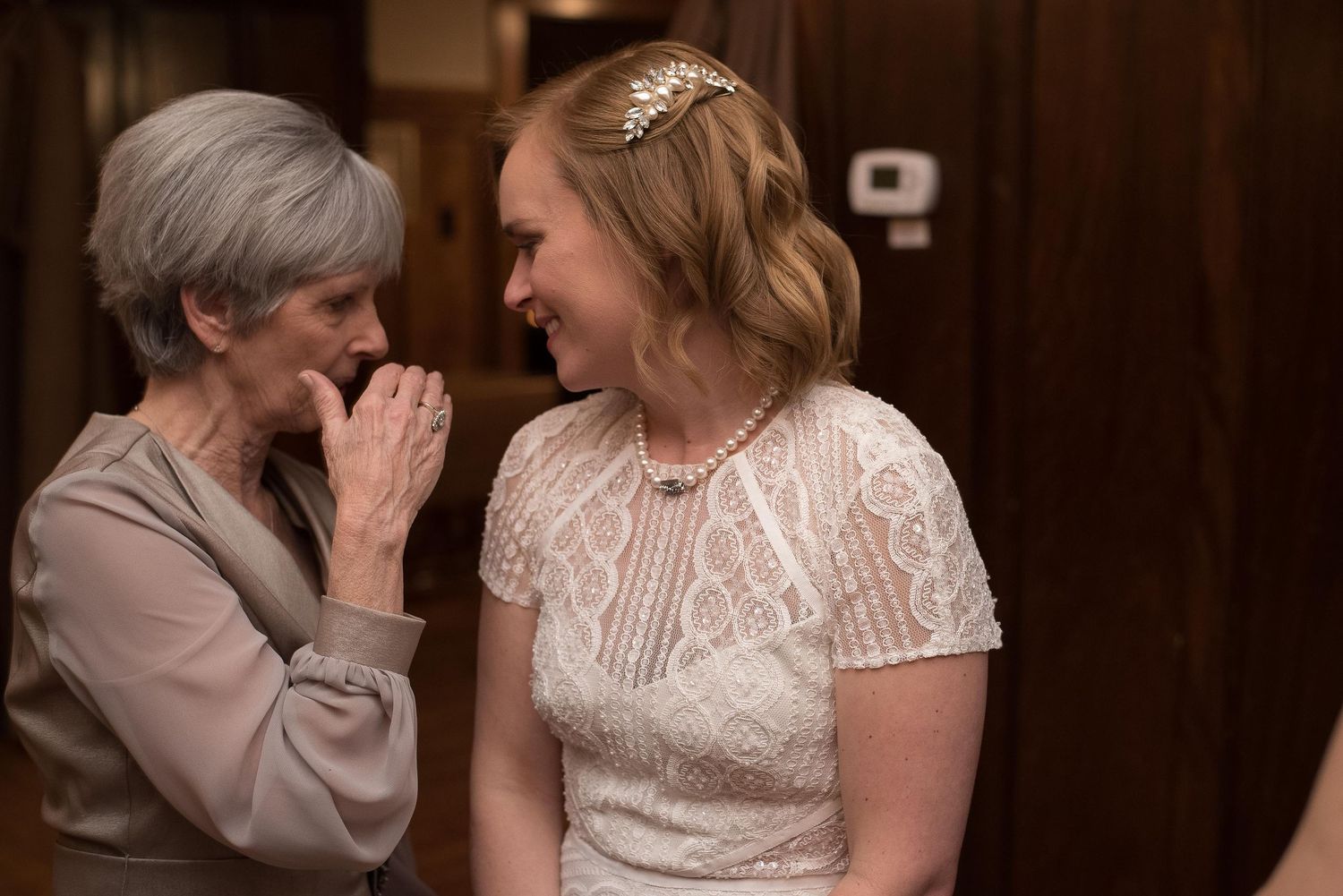 Bride in white lace dress and tiara shares a joyful moment with older woman in gray at a wedding.