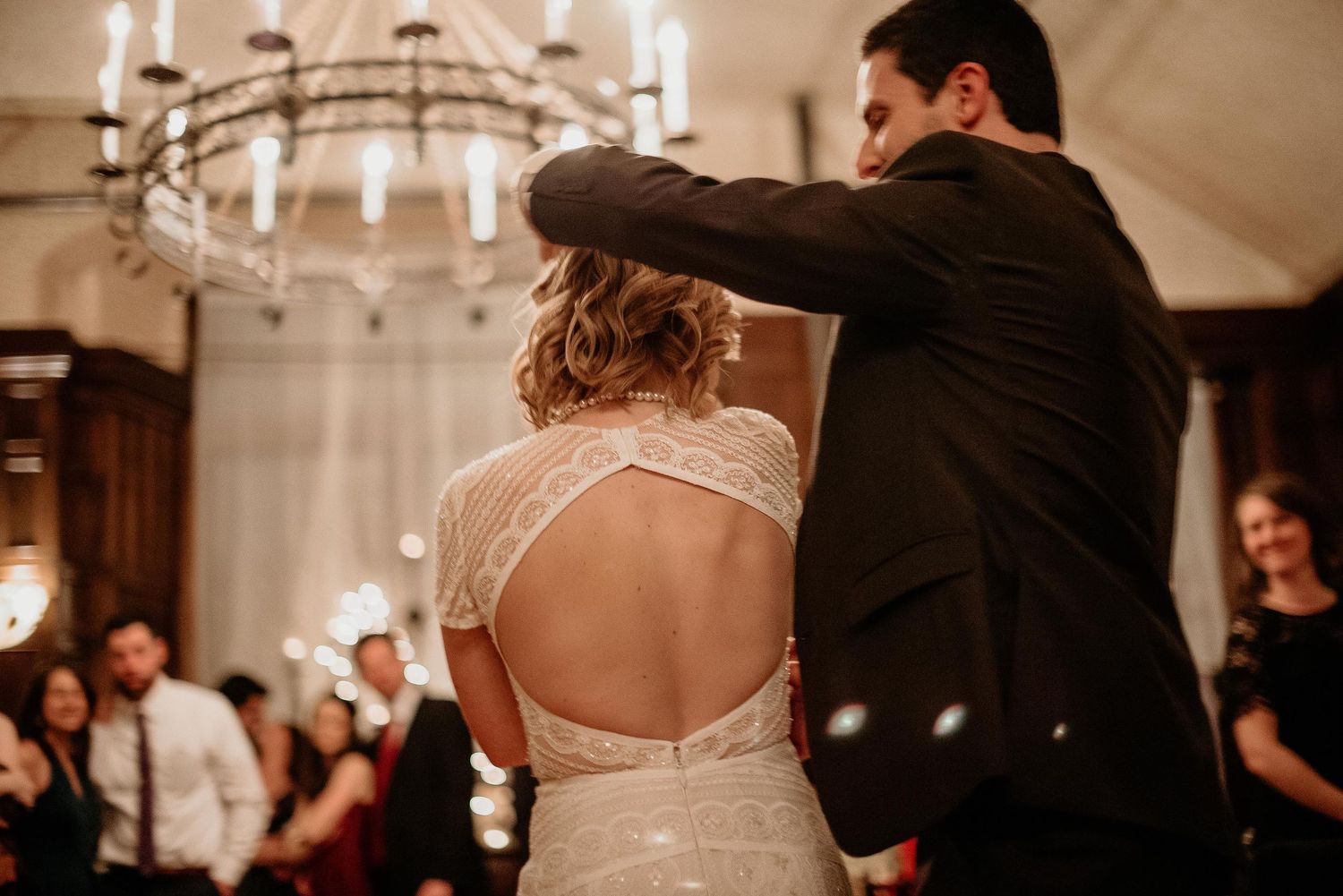 Bride and groom share first dance under chandelier in elegant wedding venue with lace backless gown.