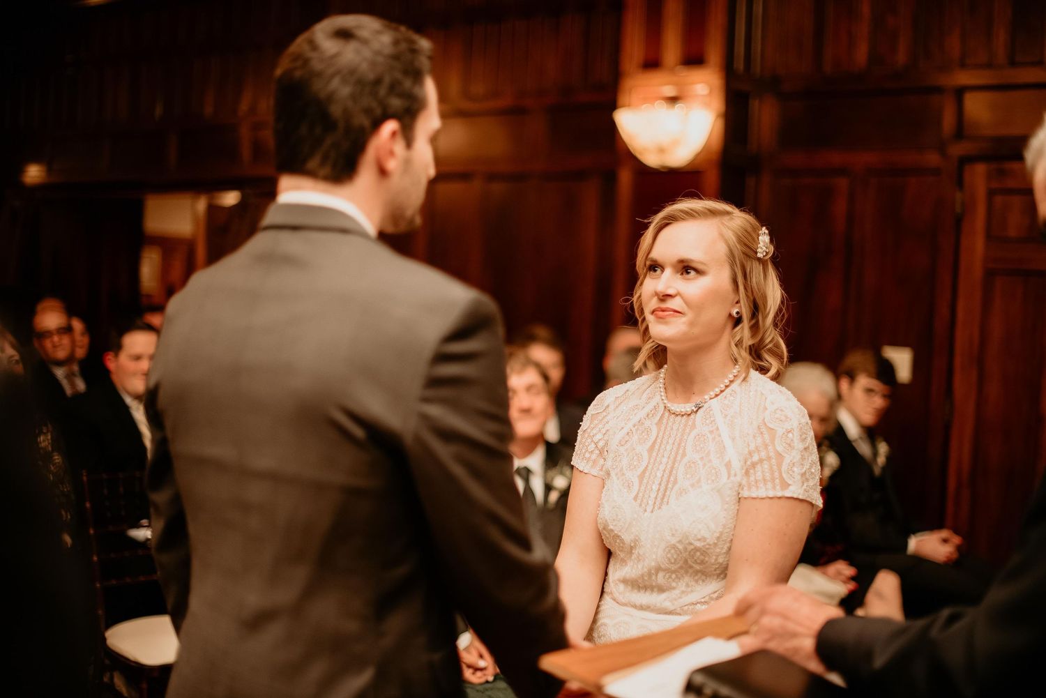 Bride smiling at groom during indoor wedding ceremony in elegant wood-paneled venue with guests seated behind.