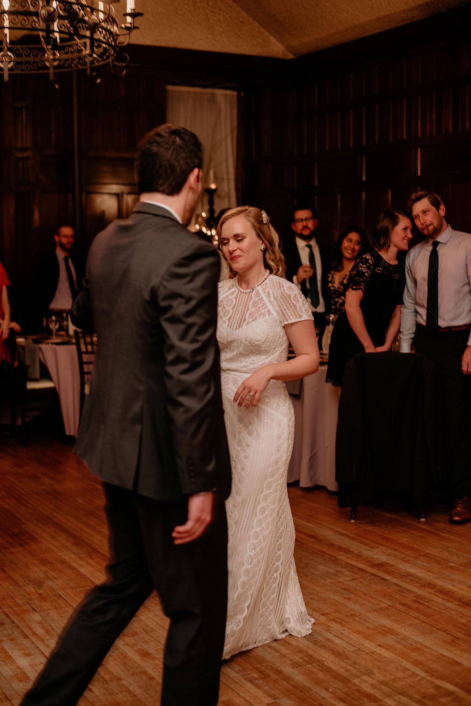 Bride in lace gown shares first dance with groom in dark suit at elegant wood-paneled wedding reception.