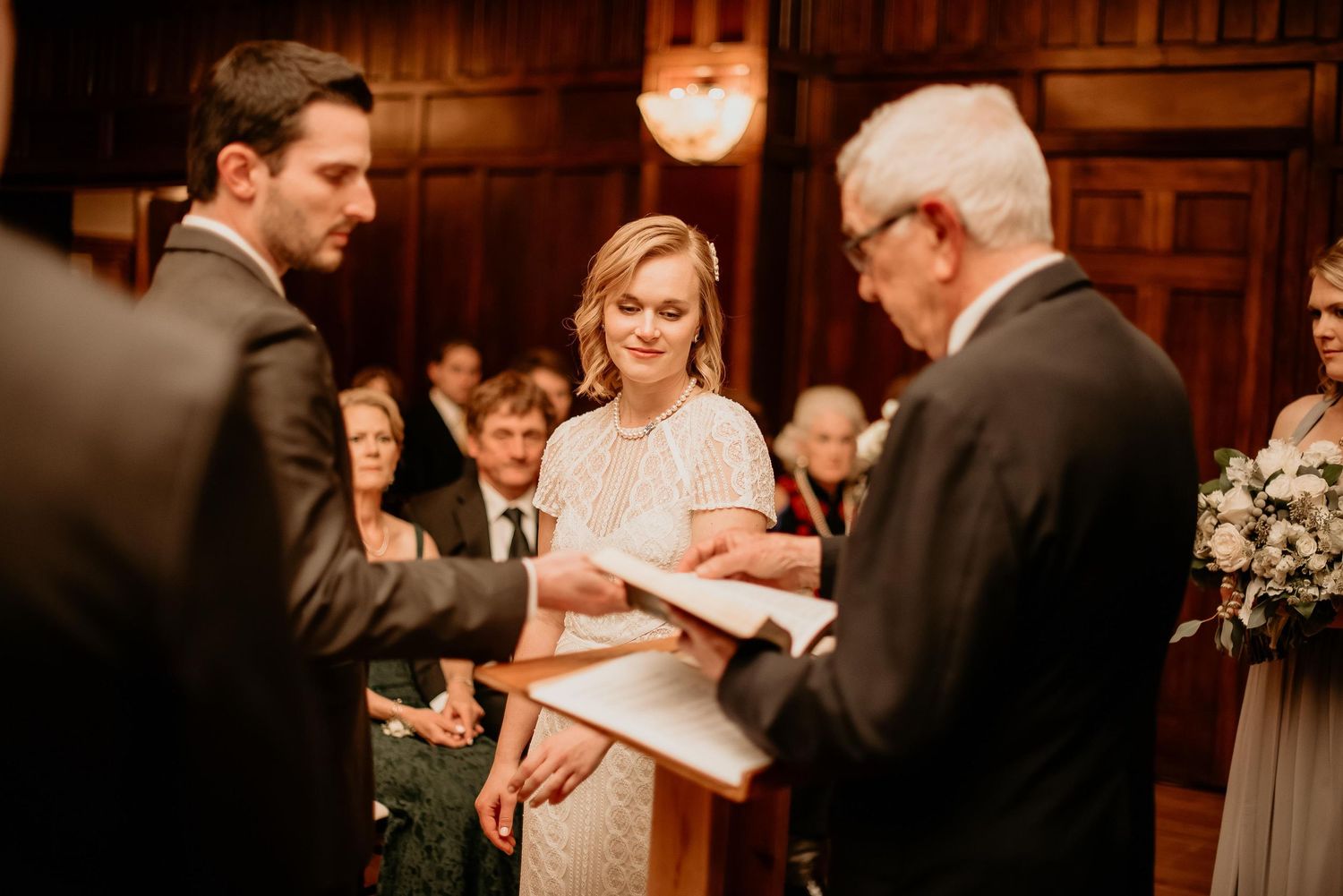 Bride and groom exchanging vows during an elegant indoor wedding ceremony with wood-paneled walls and guests seated behind.