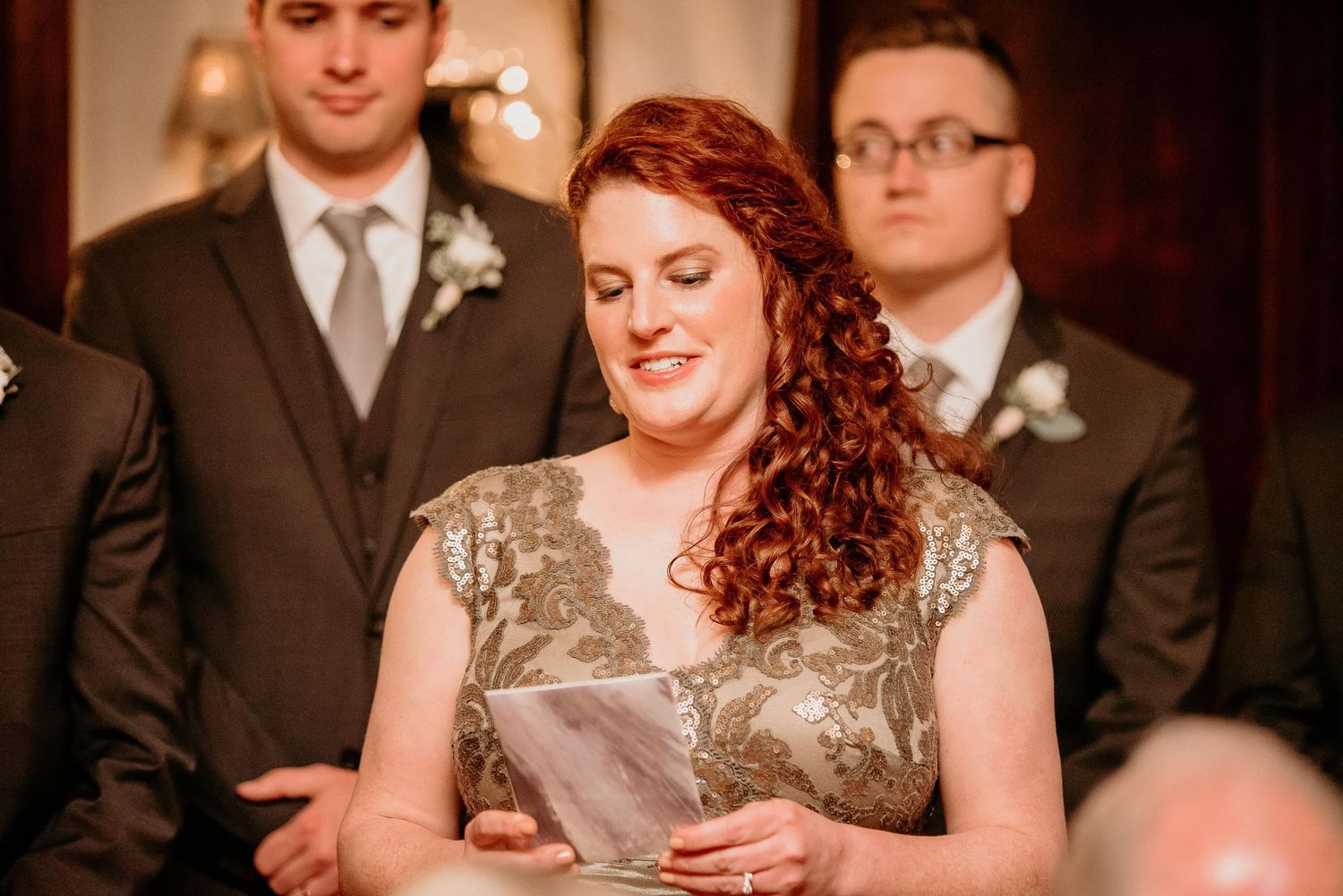Red-haired bridesmaid reads from paper during elegant wedding ceremony, with groomsmen in suits visible behind her.