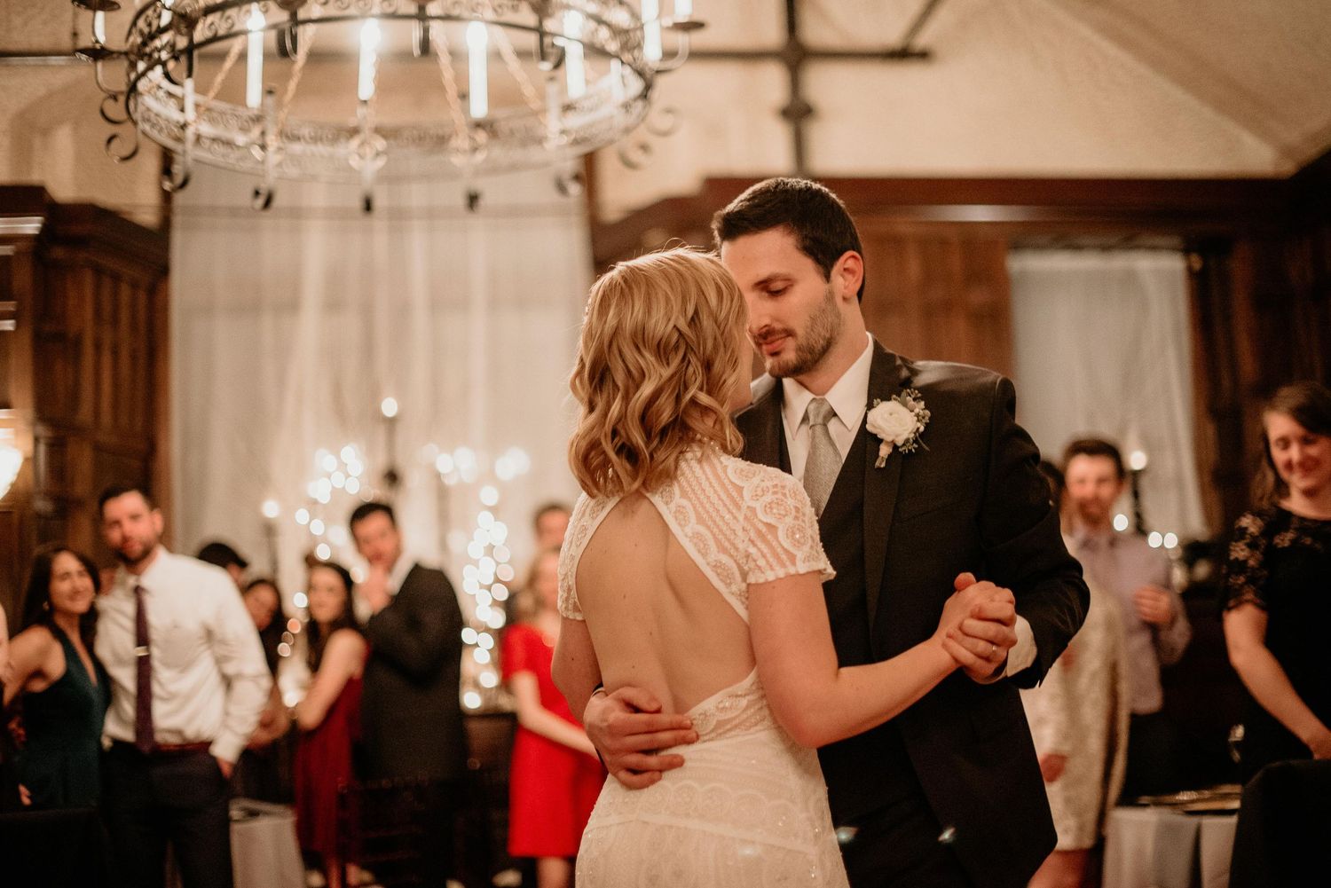 Bride and groom share their first dance at elegant wedding reception with chandelier and guests in background.