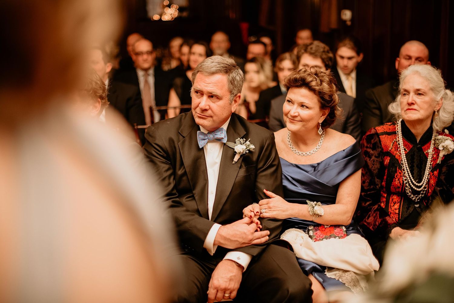 Elderly couple holds hands while seated as wedding ceremony guests, man wearing boutonniere in suit.