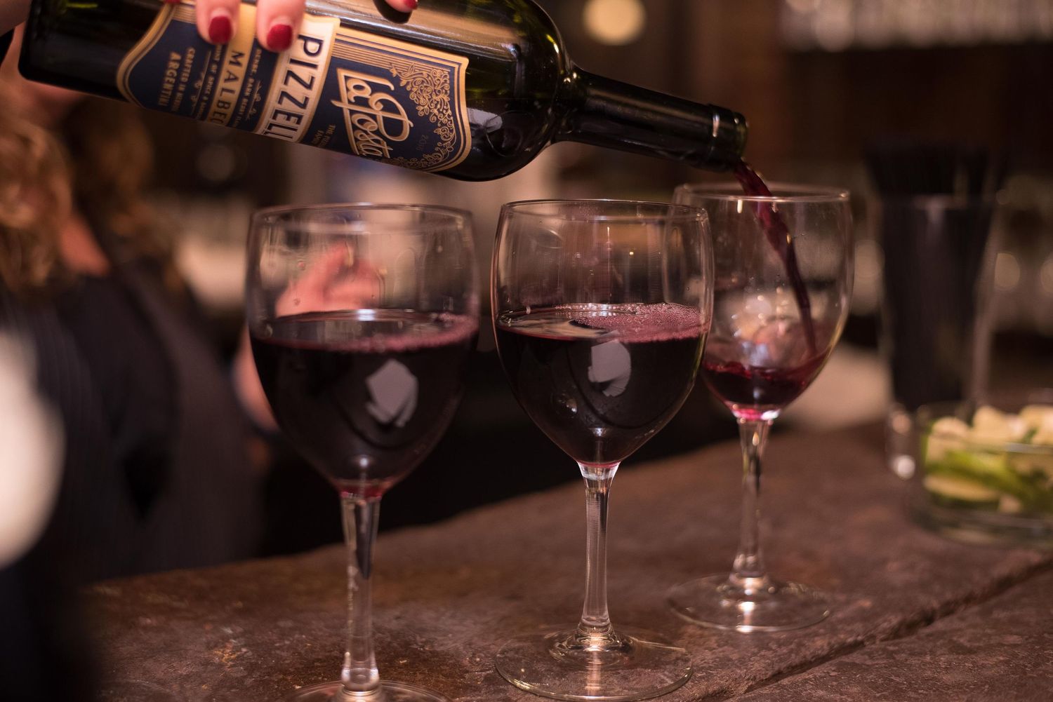 Red wine being poured into three elegant wine glasses on a rustic bar counter at a dimly lit restaurant.