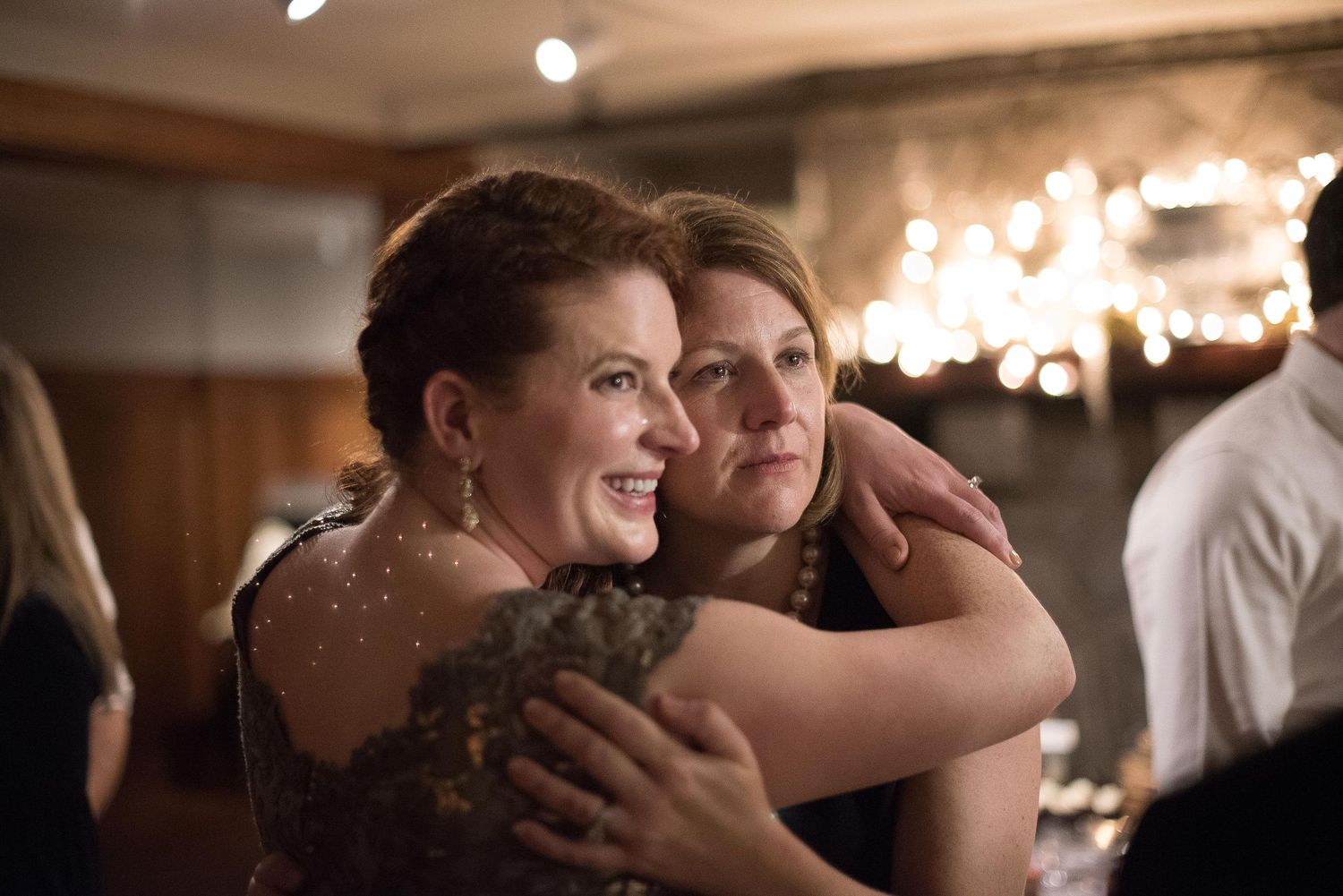 Two women share a warm embrace at an elegant indoor event, smiling together under soft ambient lighting.