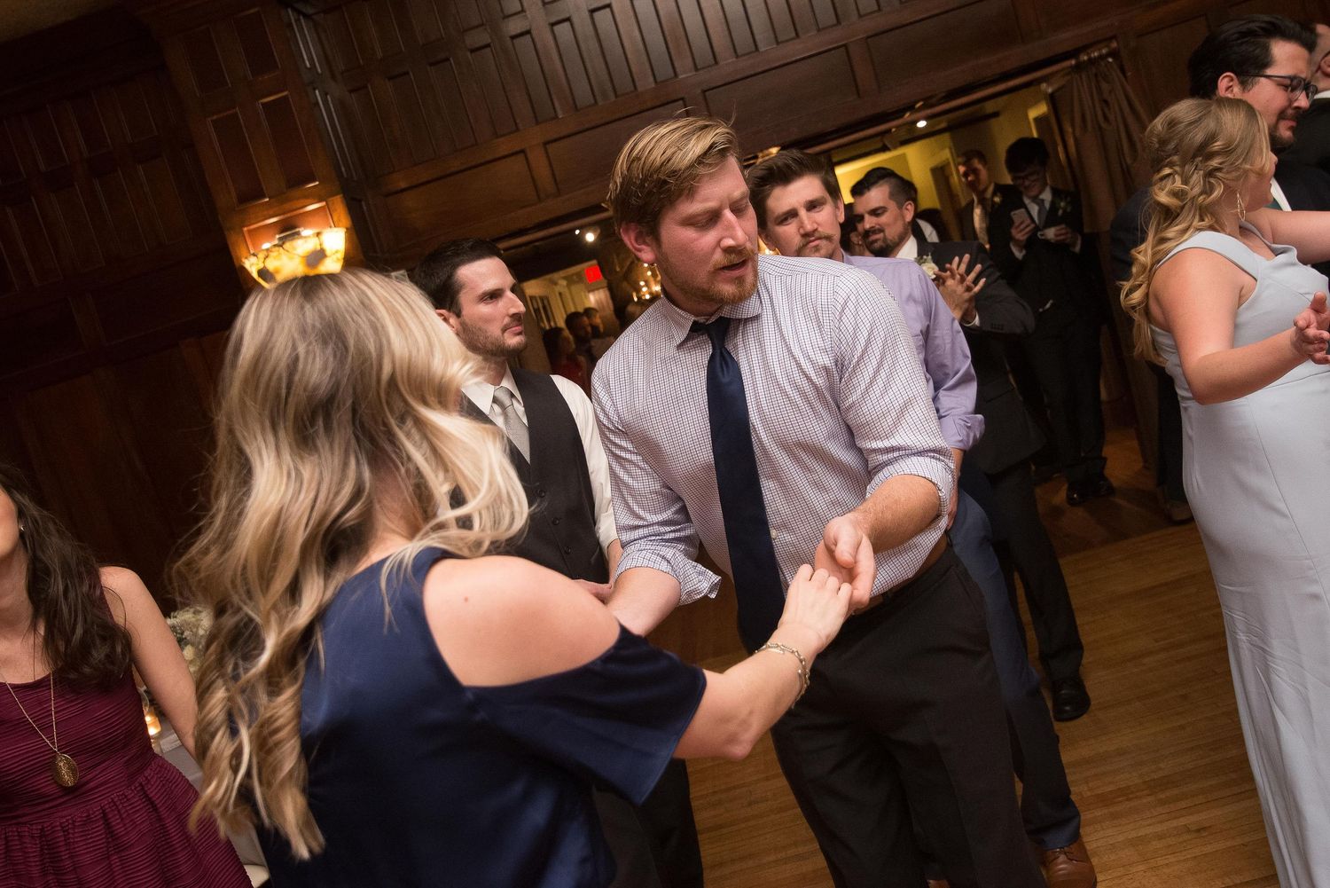 Guests dancing and holding hands at a lively indoor wedding reception in a wood-paneled ballroom.