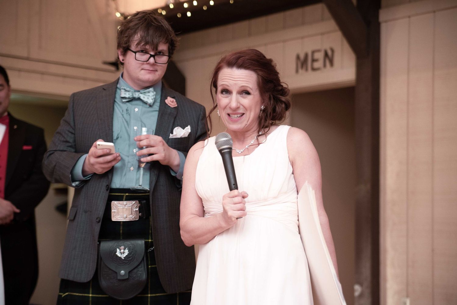 Bride in white dress holds microphone beside man in kilt and bow tie at wedding reception near MEN sign.