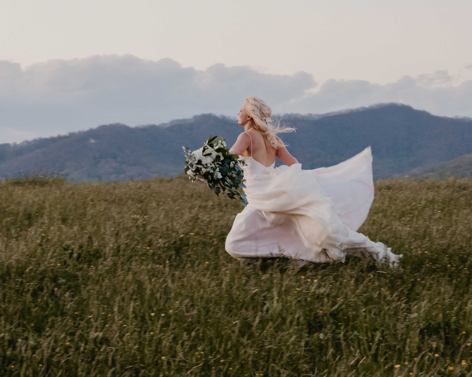 Bride in flowing white gown running through mountain meadow holding wildflower bouquet with misty hills in background.