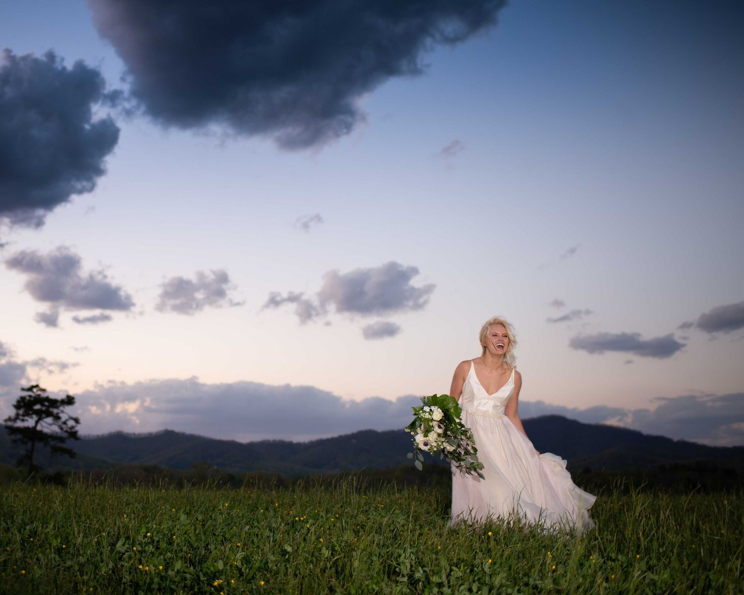Bride in white gown holding bouquet stands in green field with mountain backdrop at sunset.
