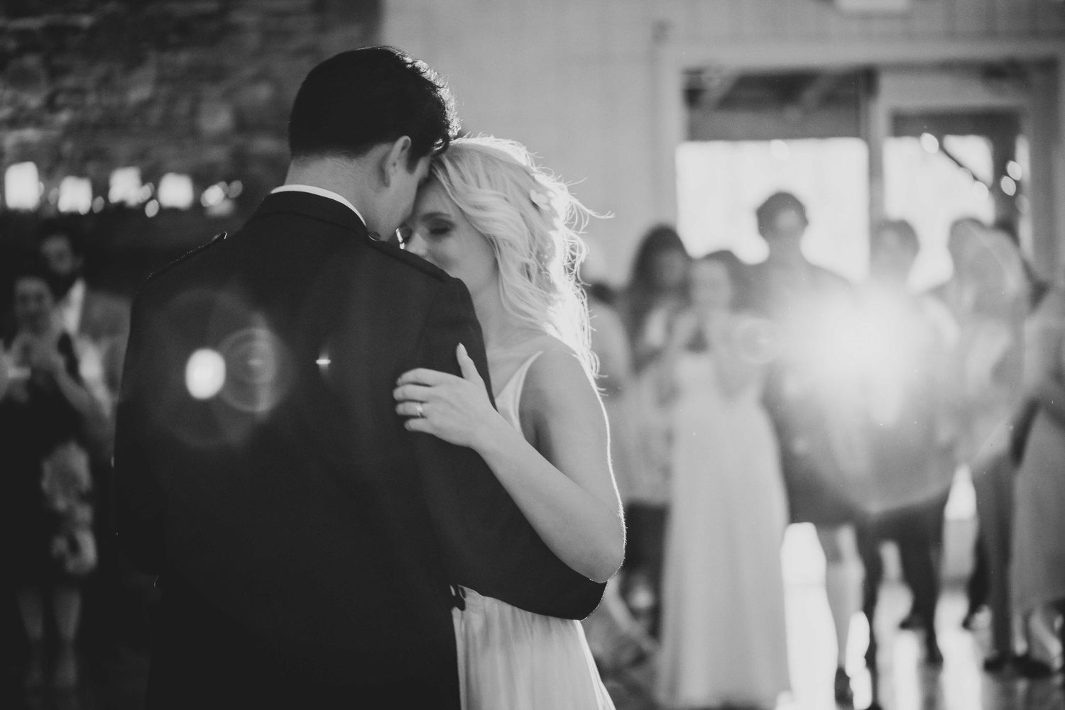 Black and white photo of a couple sharing their first dance at a wedding reception with guests watching.