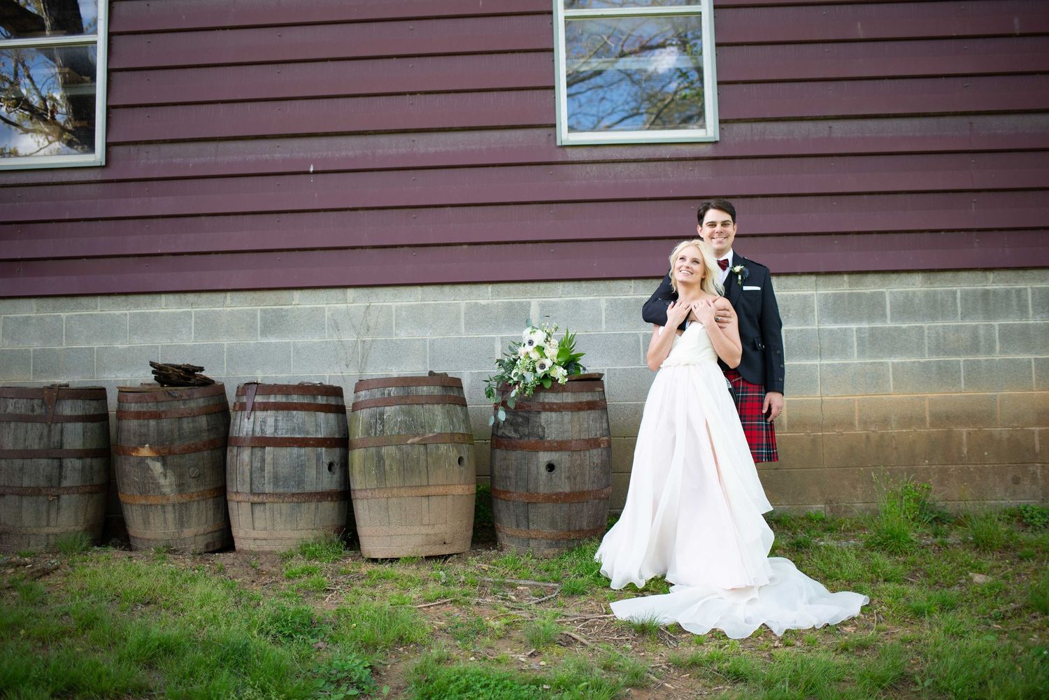 Bride and groom posing beside rustic wooden barrels outside a barn venue on their wedding day.