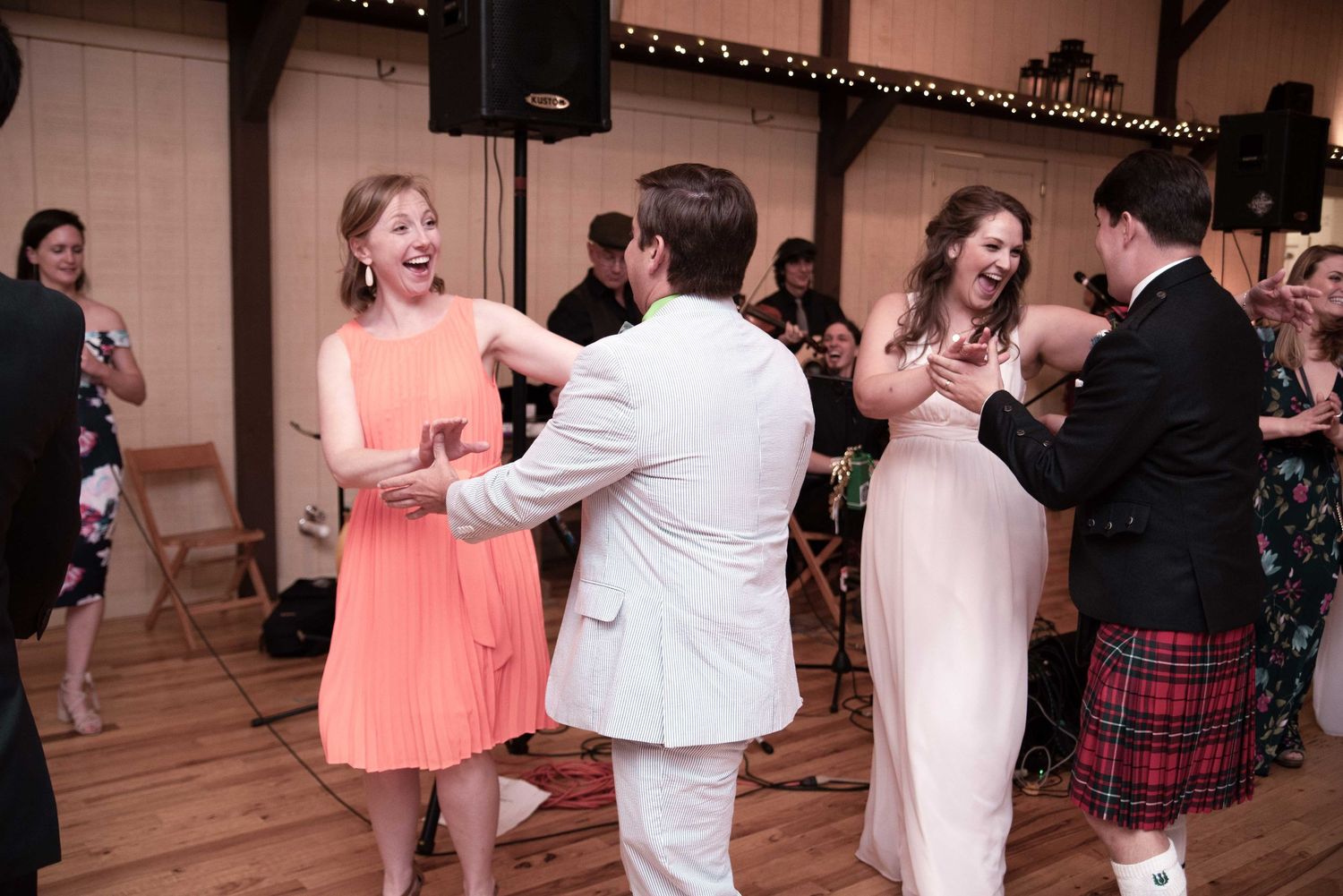 Guests dancing joyfully at a wedding reception in a rustic barn venue, wearing formal attire including a tartan kilt.