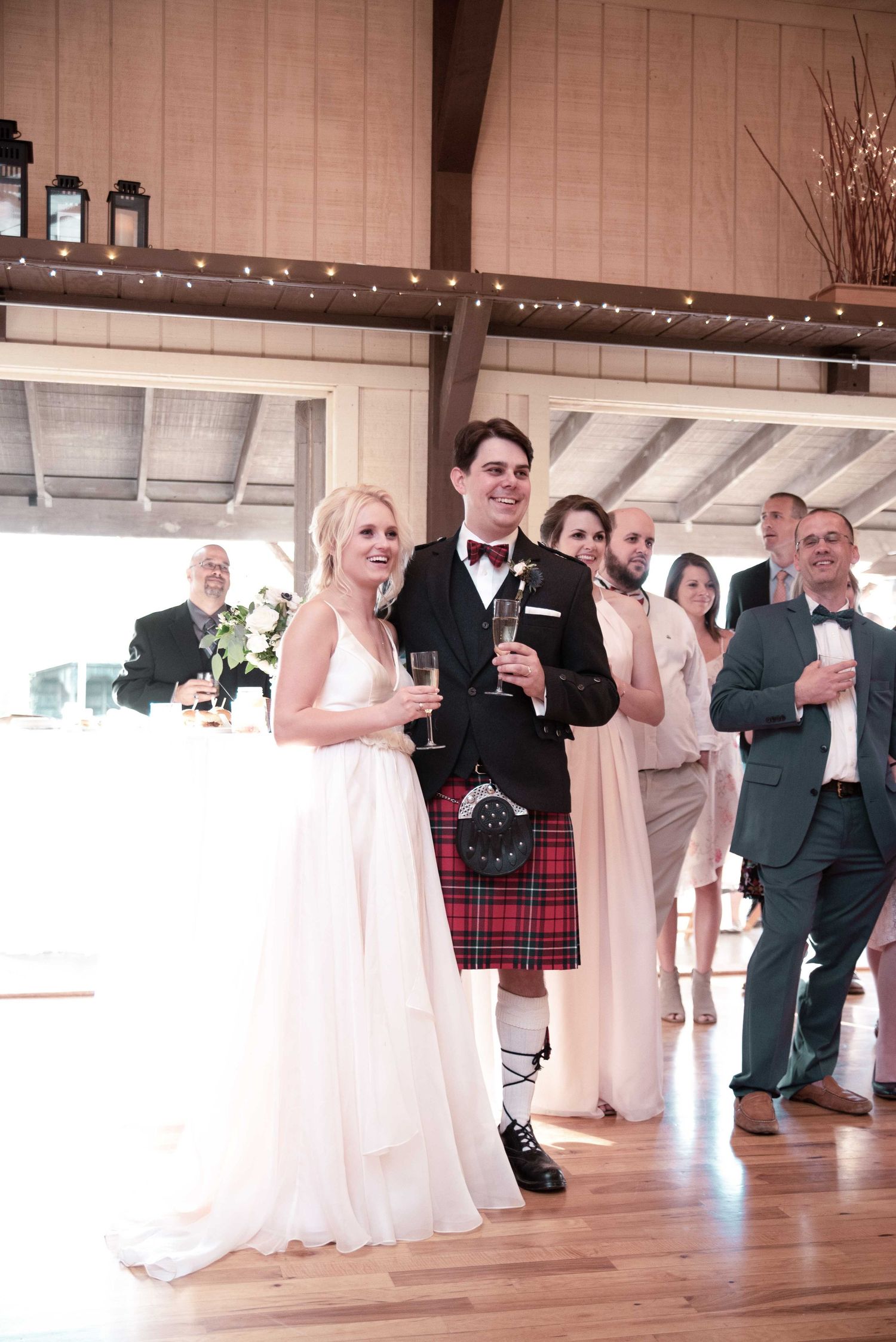 Bride in white gown and groom in Scottish kilt toast at rustic barn wedding reception with guests.