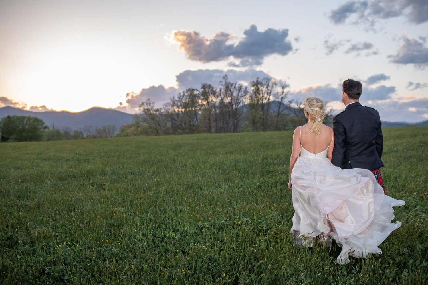 Bride and groom walking through lush green field at sunset with mountain backdrop on their wedding day.