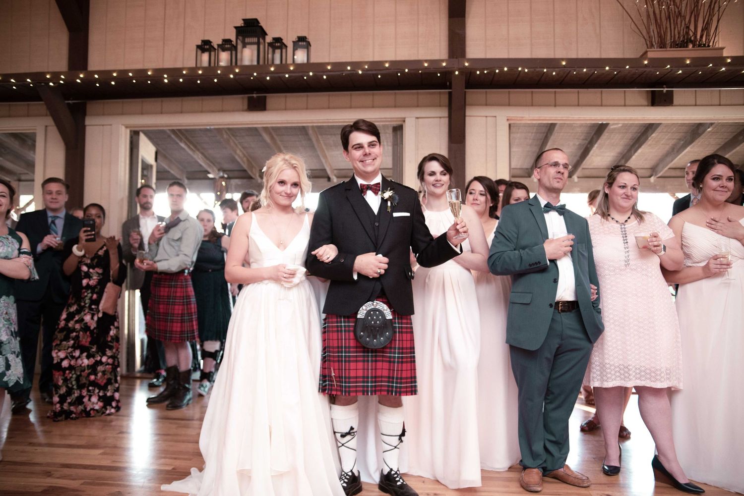 Groom in red tartan kilt and black jacket stands with bride in white gown at elegant barn wedding reception.