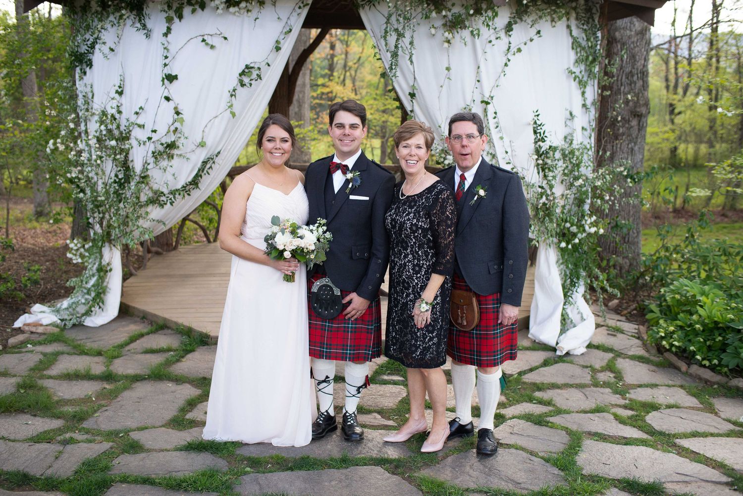 Wedding party in kilts and bride with bouquet pose under white floral arbor outdoors.
