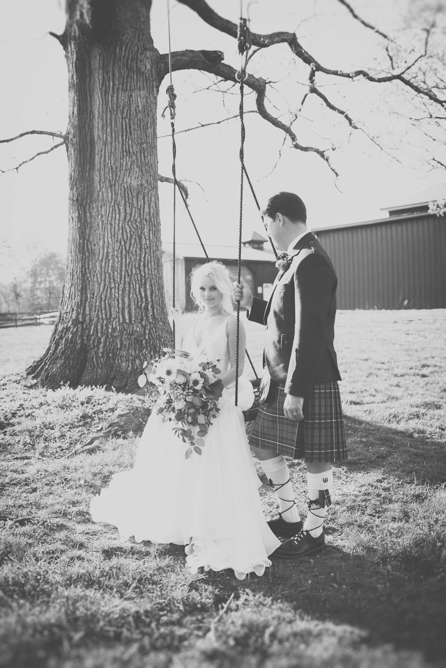 Black and white wedding photo of bride in white gown and groom in kilt standing by a tree swing outdoors.