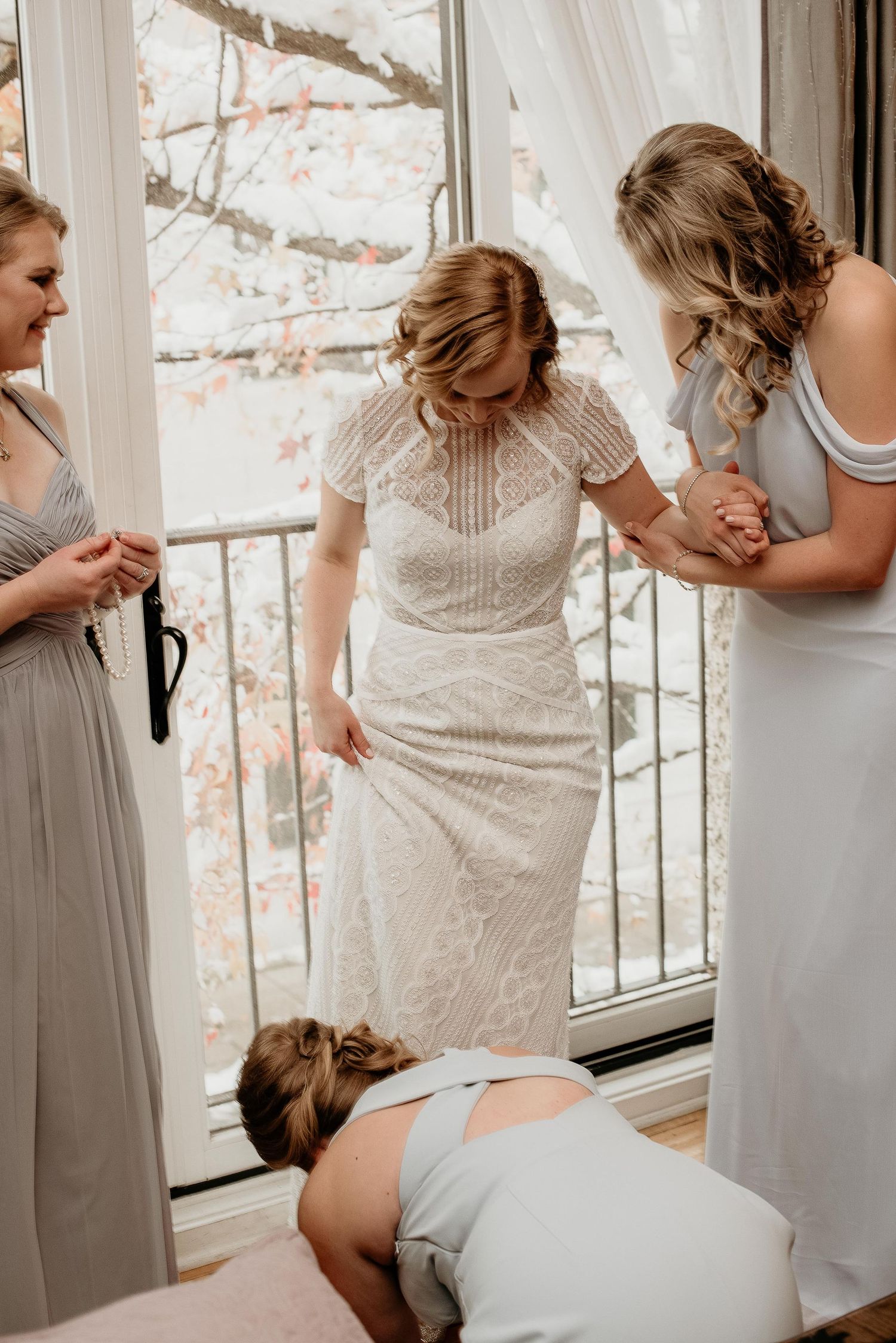 Bride in lace wedding dress being helped by bridesmaids in gray dresses near a bright window.