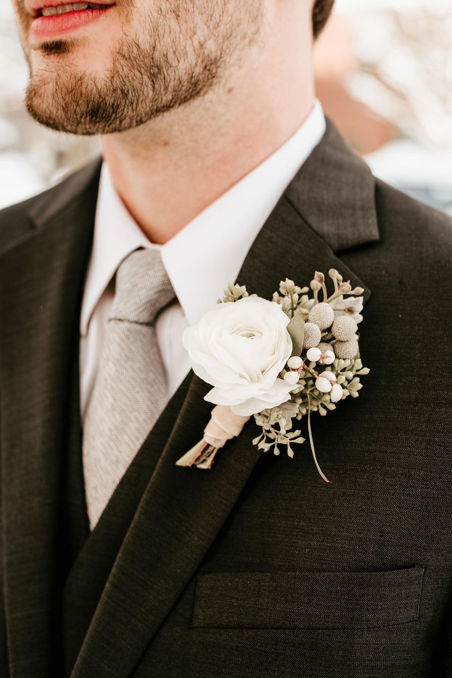 Groom wearing dark suit with white ranunculus and greenery boutonniere on lapel, paired with a neutral gray tie.