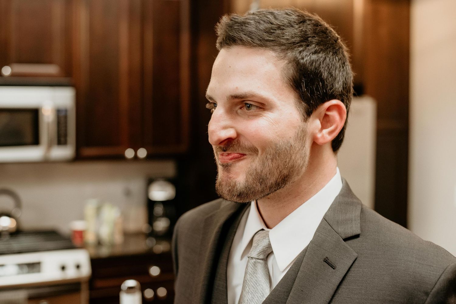 Smiling man in gray suit with tie standing in modern kitchen, dressed for formal occasion.