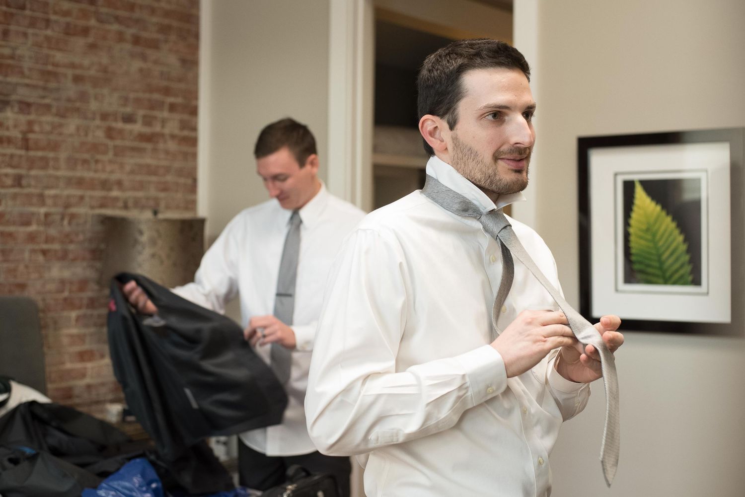Two men in white dress shirts adjusting their ties while getting ready in a room with brick walls.