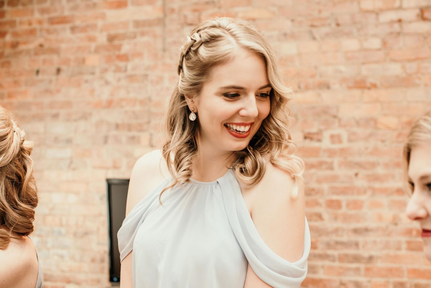 Smiling bridesmaid in cold-shoulder dress with pearl earrings and braided updo against brick wall.