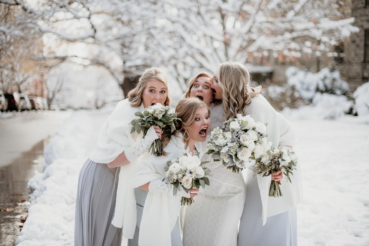 Bride and bridesmaids in white gowns holding floral bouquets laughing together in a snowy winter wedding outdoor setting.