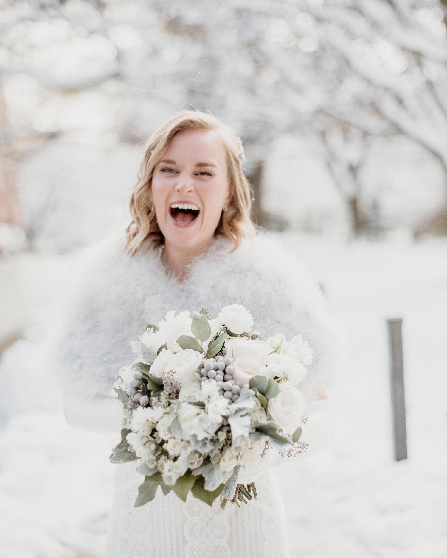 Joyful bride in white fur wrap holding elegant white floral bouquet outdoors in snowy winter setting.