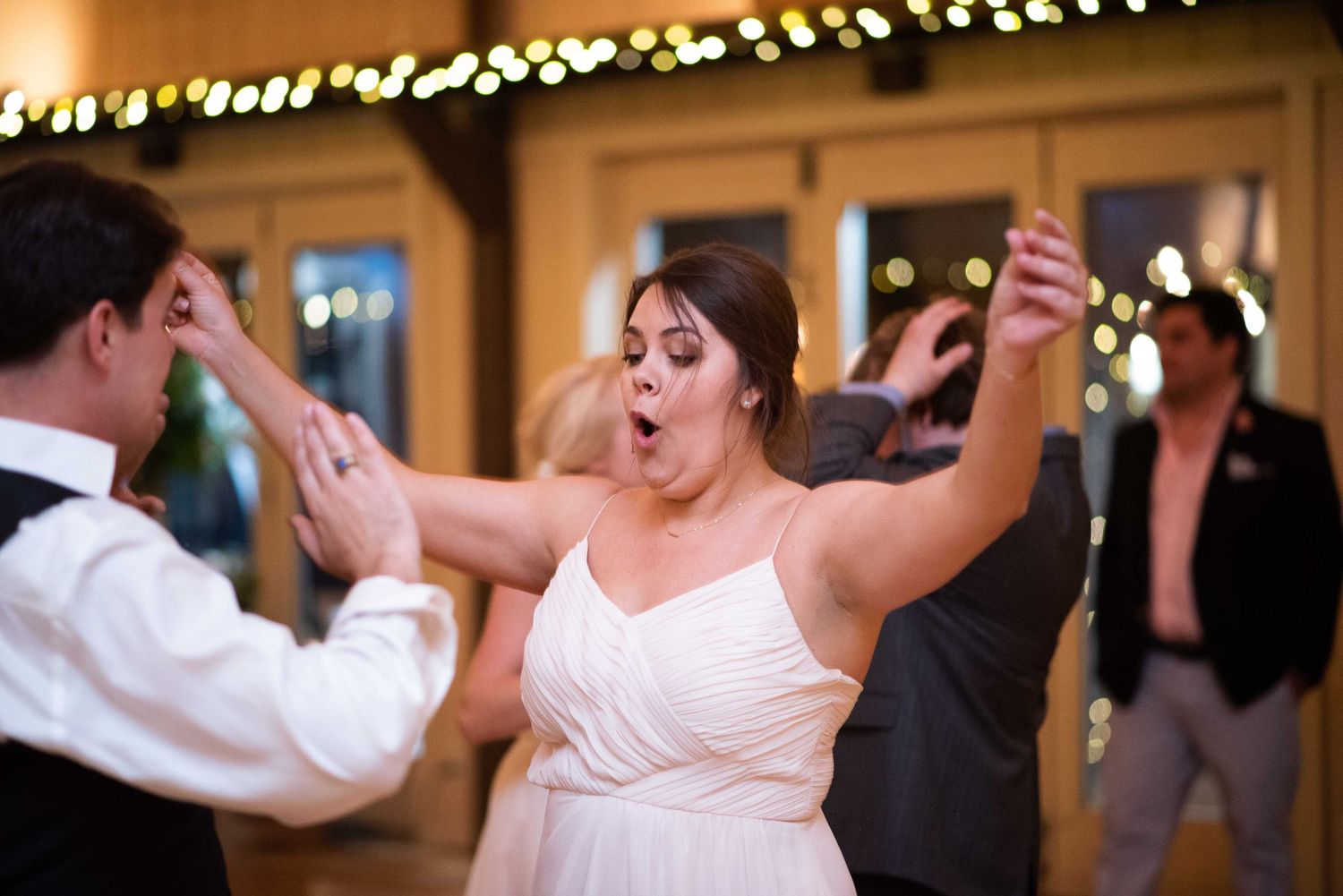 Bride in white dress dancing energetically with arms raised at wedding reception with string lights.