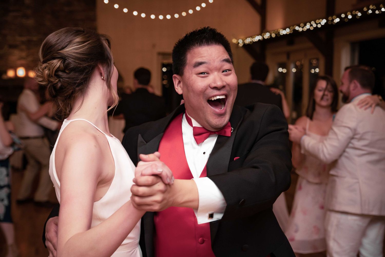 Joyful groom in black tuxedo with red vest dancing enthusiastically with bride at elegant wedding reception.